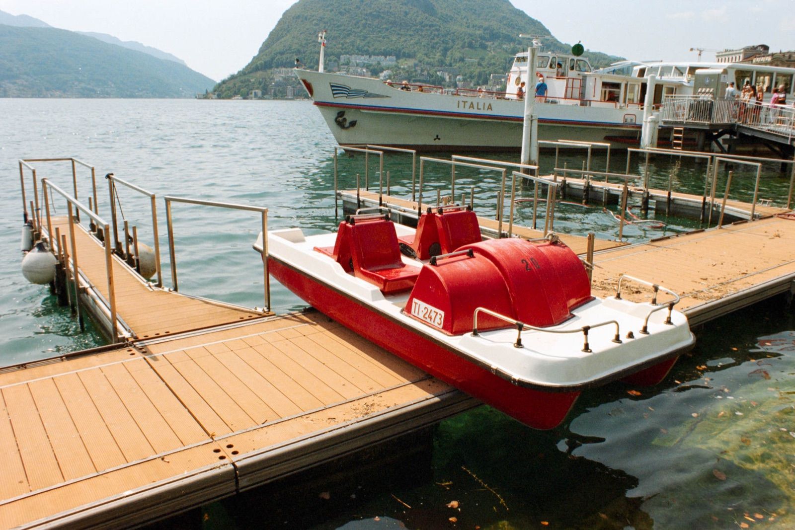 Rotes Tretboot an einem Holzsteg am See, dahinter ein weißes Passagierschiff namens Italia und ein bewaldeter Berg