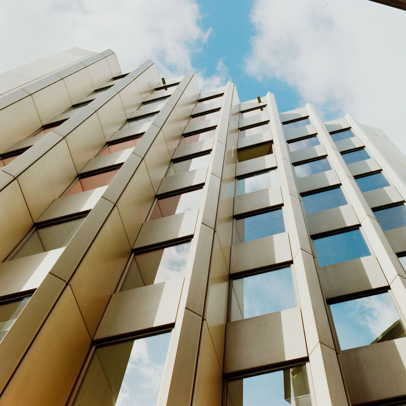 Helle, gefaltete Hochhausfassade aus steiler Untersicht, in den Fenstern spiegelt sich blauer Himmel